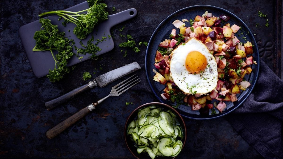 Pytt i Panna - schwedische Bratkartoffeln auf einem dunkelblauen Teller auf einem dunkelblauen Geschirrtuch auf einem dunkelblauen Hintergrund. Unten eine Schüssel mit Gurkensalat und Besteck. Links oben ein kleine Schneidebrett mit Petersilie.