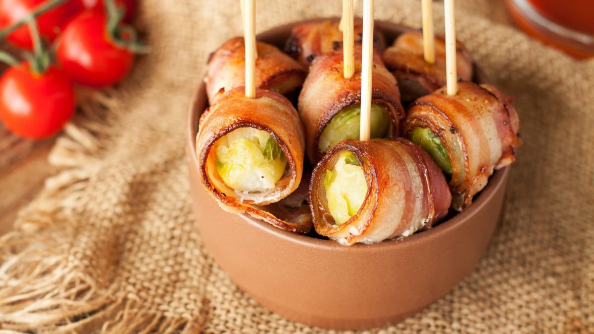 Draufsicht, schräg: Eine Schale aus Ton mit Rosenkohl-Tapas, die mit Zahnstochern fixiert sind. Die steht auf einem grobem Leintuch. Im Hintergrund liegen frische Tomaten.