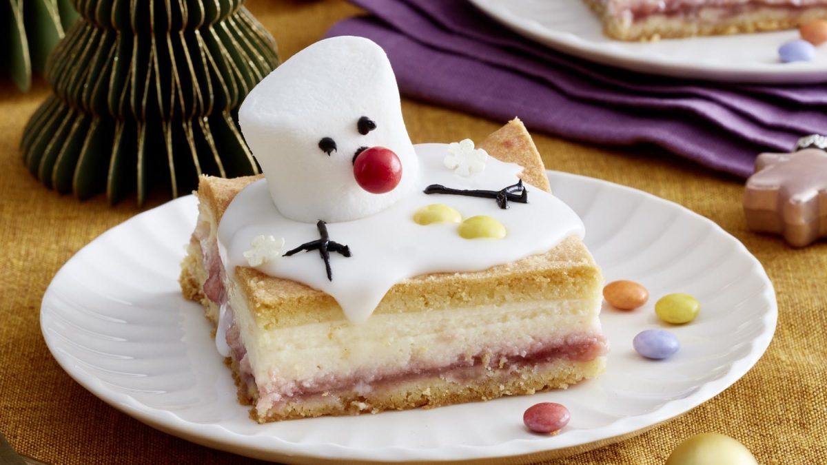 Ein Stück Schneemann-Käsekuchen auf einem weißen Teller auf einem dunkelgelben Hintergrund. Rechts oben ein violettes Geschirrtuch und ein weiterer Teller.