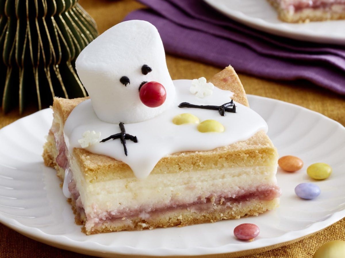 Ein Stück Schneemann-Käsekuchen auf einem weißen Teller auf einem dunkelgelben Hintergrund. Rechts oben ein violettes Geschirrtuch und ein weiterer Teller.