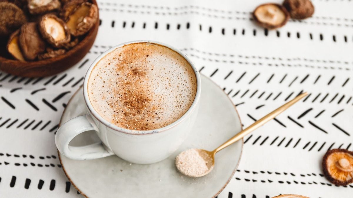 Eine weiße Tasse mit dem Shiitake-Latte auf einem weißen Tuch mit schwarzem Muster. Daneben links noch eine Schale mit getrockneten Shiitake-Pilzen.