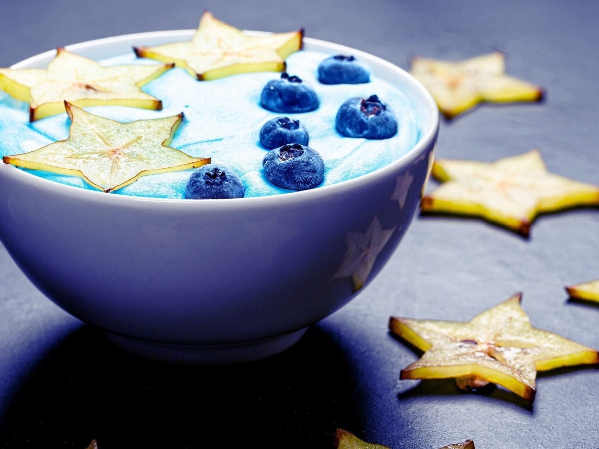 Frontal: eine weiße Schüssel mit einer Sternfrucht-Blaubeeren-Bowl auf blauem Untergrund. Drum herum liegen Scheiben einer Sternfrucht.