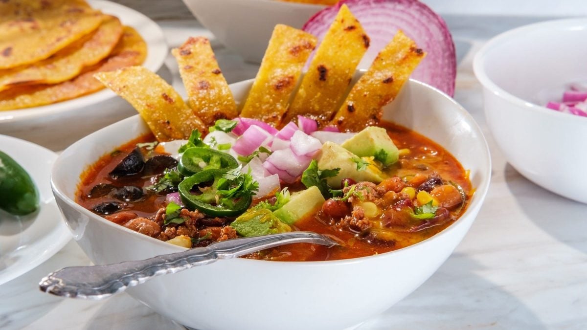 Eine weiße Schüssel mit der Taco-Suppe auf weißem Marmor. In der Suppe ein Löffel und etwas Mais-Fladenbrot. Alles von der Seite fotografiert.