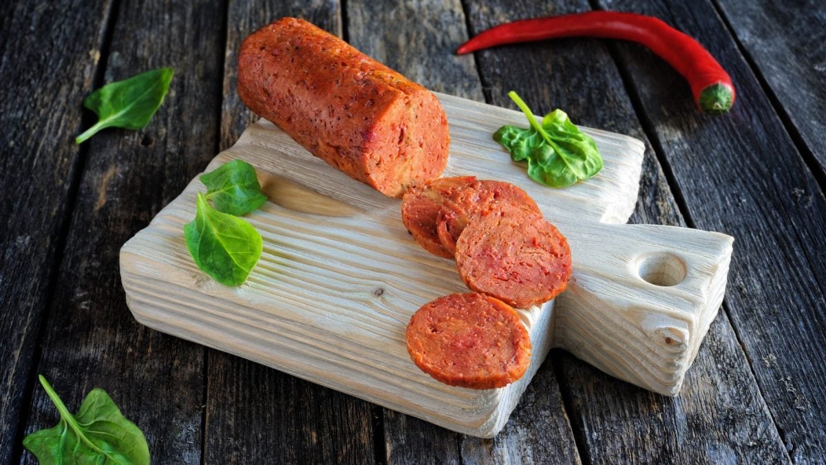 Vegane Salami auf einem Schneidebrett auf einem hölzernen Hintergrund. Oben auf dem Foto eine rote Chili. Auf dem Schneidebrett und dem Hintergrund Basilikumblätter verteilt.