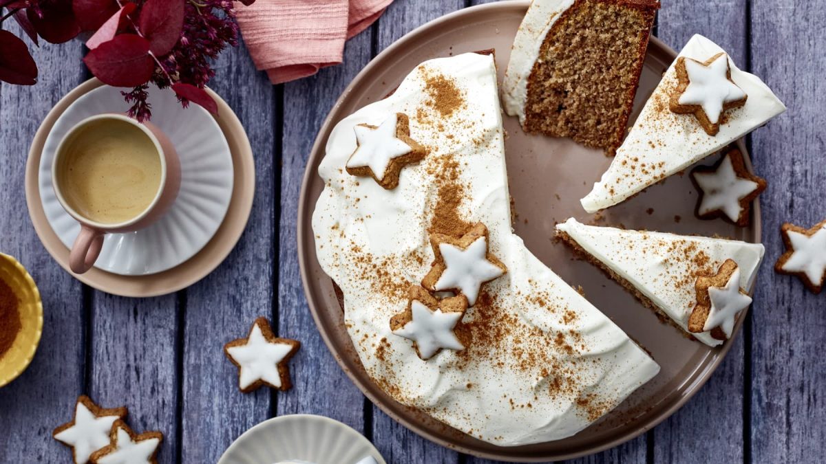 Ein Zimtstern-Nusskuchen auf einem braunen Teller auf einem dunkelgrauen Hintergrund. Links eine Tasse Kaffee und Blumen. Auf dem Hintergrund Zimtsterne verteilt.