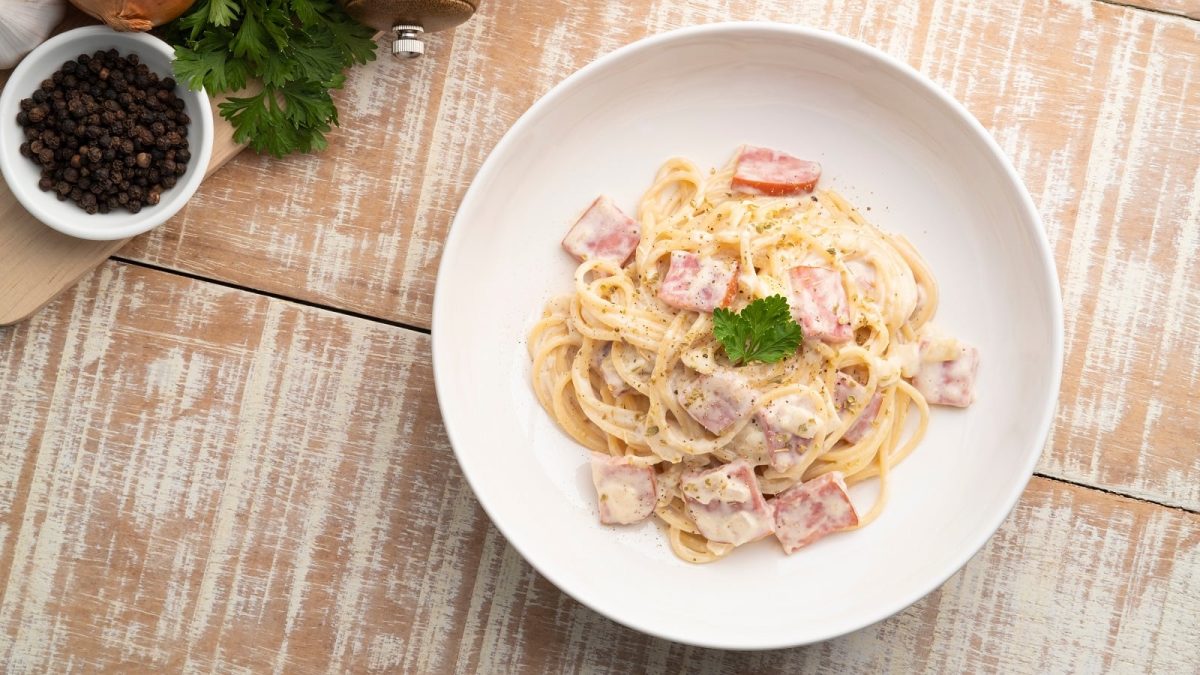Ein weißer Teller mit Spaghetti mit Schinkensahnesauce auf einem hellbraunen Holztisch.