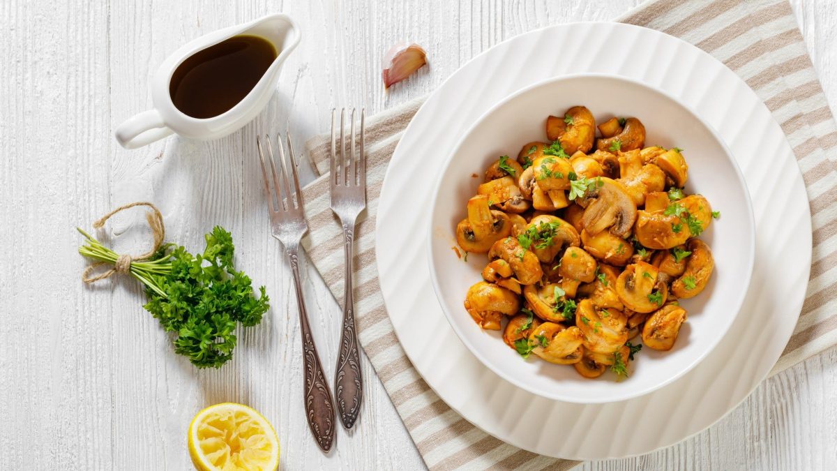 Spanische Champiñones al jerez – Knoblauch-Champignons – auf weißem Teller. Daneben Gabeln, Zitrone, Petersilie und eine Knoblauchzehe.