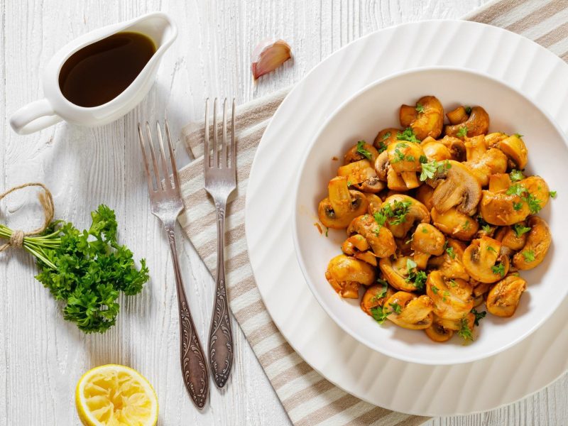 Spanische Champiñones al jerez – Knoblauch-Champignons – auf weißem Teller. Daneben Gabeln, Zitrone, Petersilie und eine Knoblauchzehe.