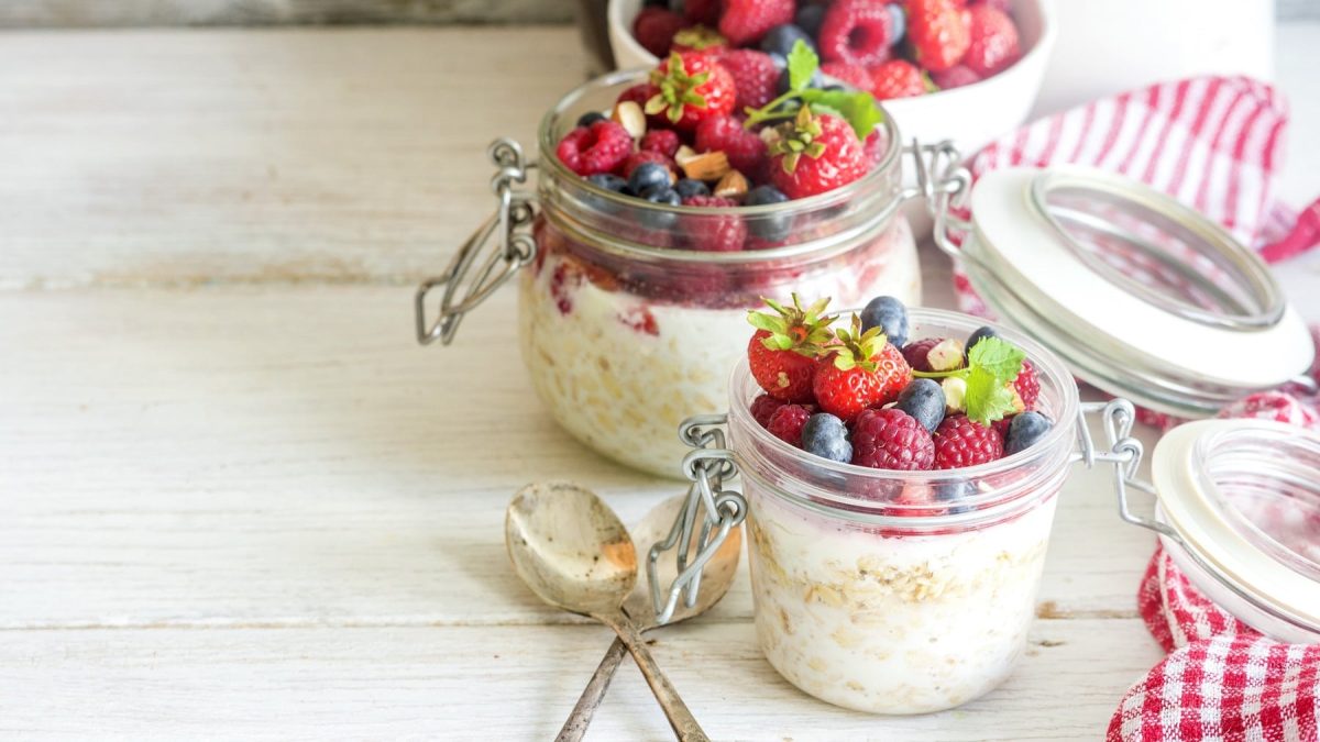 Zwei Weckgläser mit der Cheesecake Bowl und einer Menge Früchten auf einem hellen Holztisch von der Seite fotografiert. Daneben rot-weiß-karierte Tischdecken.