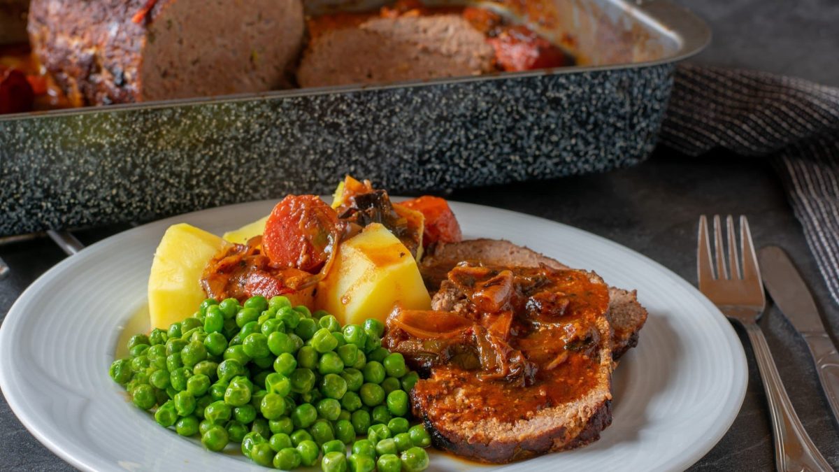 Draufsicht, schräg: Ein weißer Teller mit Omas Hackbraten mit Sauce und Erbsen steht auf einem Holztisch. Im Hintergrund ist der Hackbraten in einer Kastenform.