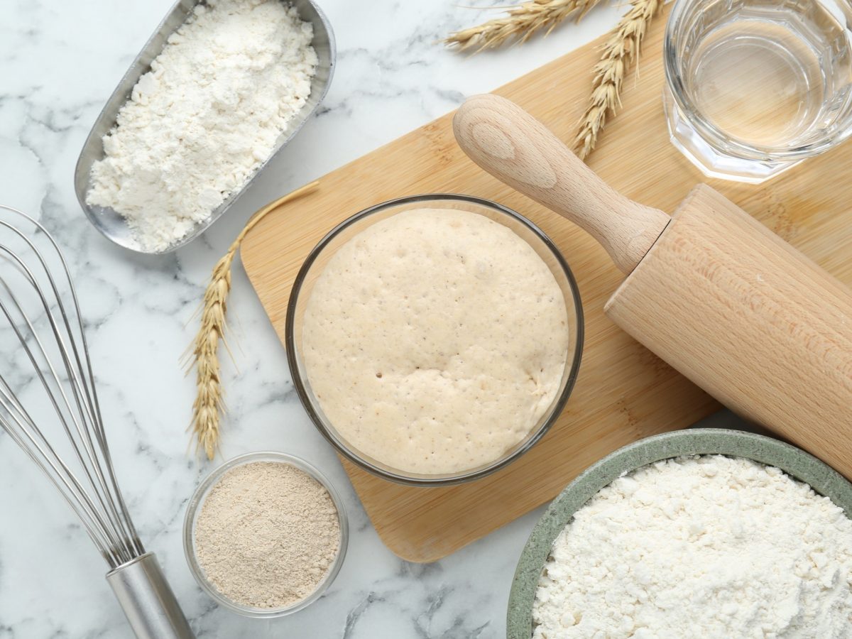 Draufblick: Self Rising Flour in einer Holzschale, Hefeteig in einer Schale, eine Holzrolle um Teig auszurollen, ein Schneebesen aus Metall / heller Untergrund
