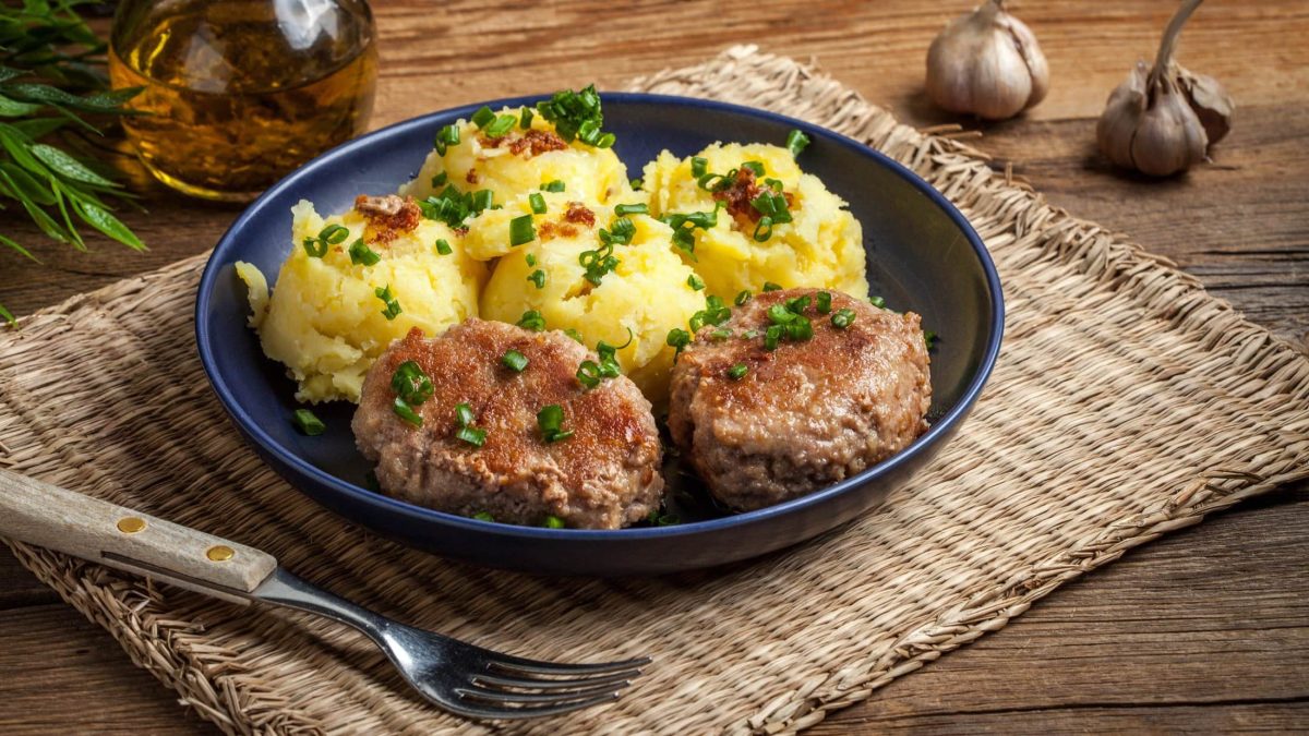 Draufsicht, schräg: Ein Teller mich Kartoffelpüree und n, beziehungsweise österreichische Hackbällchen steht auf einer Bastunterlage auf einem Tisch. Daneben liegen eine Gabel und zwei Knoblauchzehen.