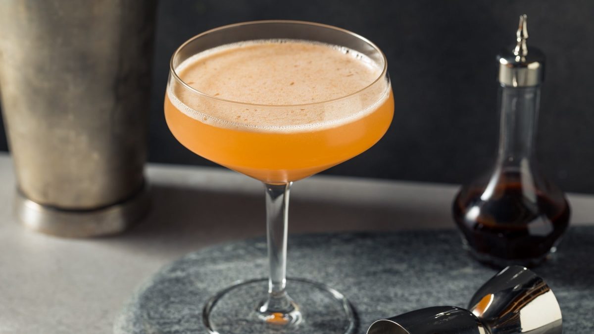 Ein Martiniglas mit dem Golden Velvet auf dunklem Unter- und Hintergrund. Daneben ein Barmaß und eine Flasche mit dem Zimtsirup. Alles von der Seite fotografiert.