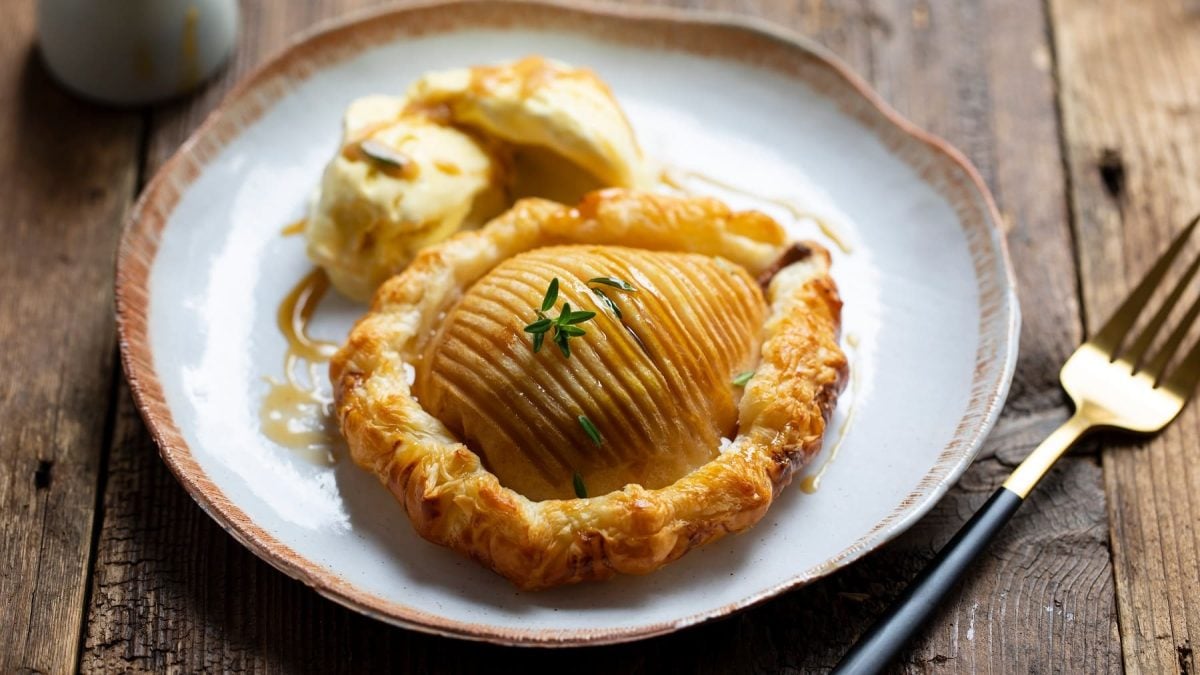 Die Hasselback-Birne in Blätterteig gebacken auf einem grauen Teller mit Eiscreme. Alles von schräg oben auf einem dunklen Holztisch fotografiert.
