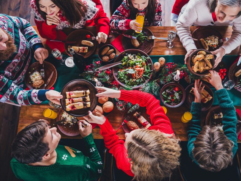 Draufsicht auf einen Tisch, der mit Essen für Heiligabend eingedeckt ist und an dem eine Familie mit Weihnachtspullovern sitzt.