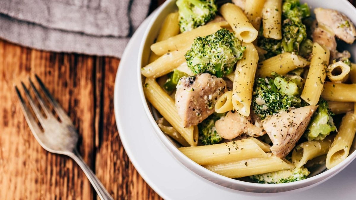 Draufsicht: Eine Schüssel Hühnchen-Pasta, die auf einem Teller steht. Daneben liegen eine Gabel und ein graues Tuch.