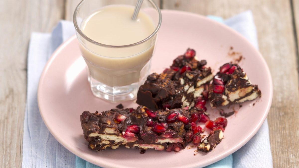 Eine Portion Kalter Hund-Kuchen mit Granatapfelkernen und ein Glas Milch auf einem rosa Teller. Der Teller steht auf einem hellblauen Geschirrtuch und der Hintergrund ist hölzern.