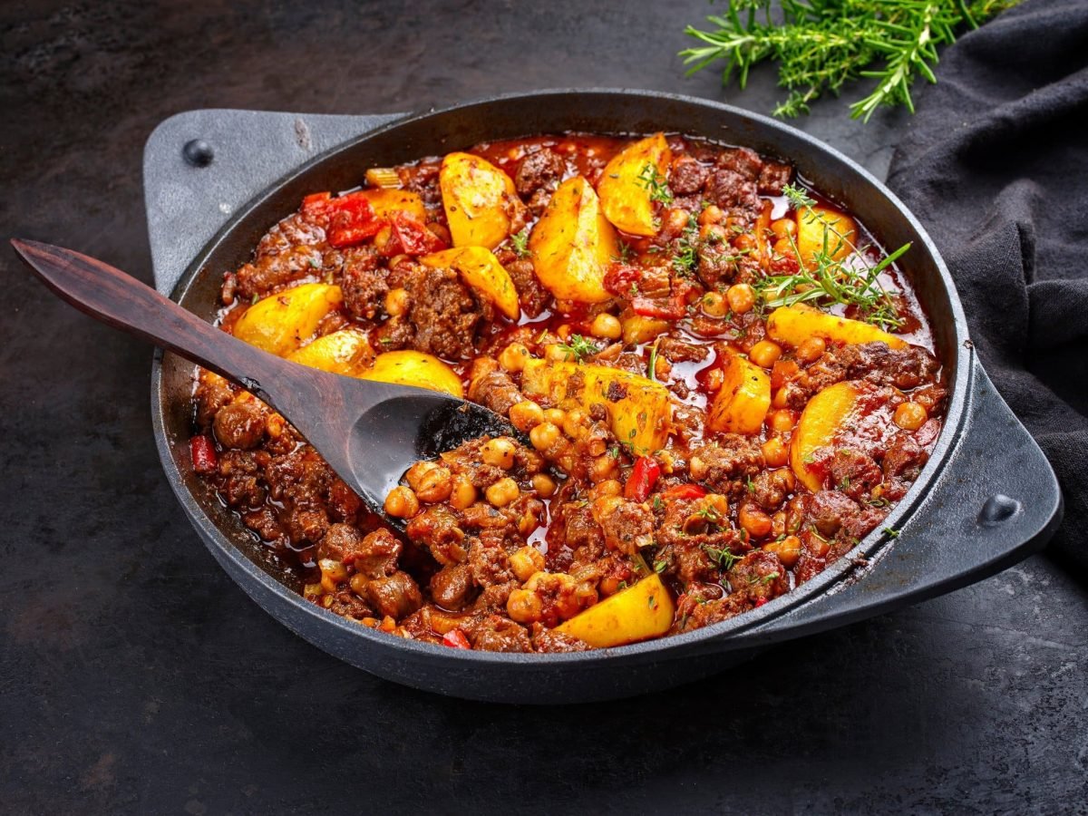 Draufsicht, schräg: eine Italienische Hackfleisch-Pfanne mit Kartoffeln steht auf einem schwarzen Untergrund. In der Pfanne ist ein Holzlöffel. Im Hintergrund ist frischer Rosmarin.