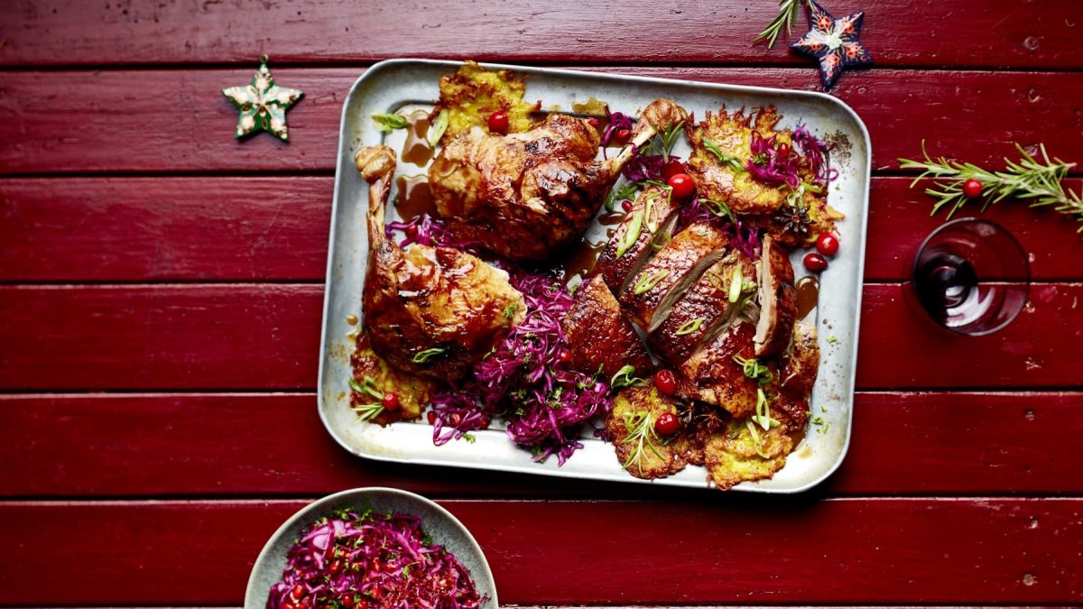 Kanton-Ente mit Rösti und spicy Rotkrautsalat auf einem Backblech auf einem roten, hölzernen Hintergrund. Auf dem Hintergrund sind zwei Sterne als Weihnachtsdekoration und ein Mistelzweig verteilt.