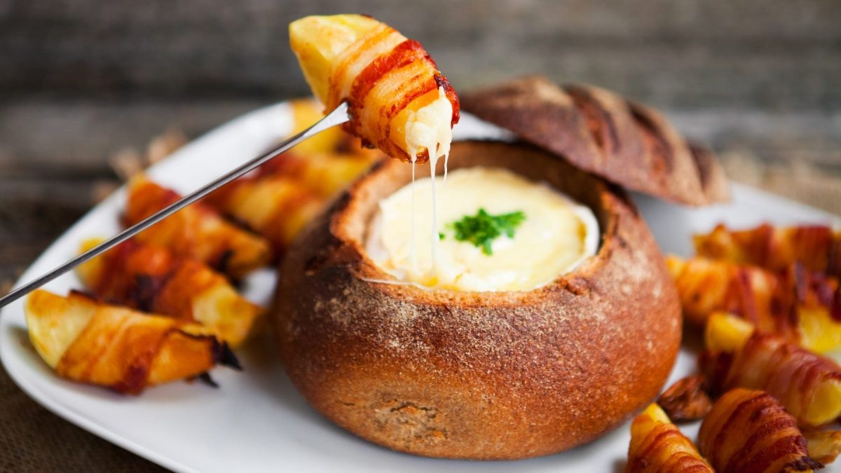 Das Fondue in einem ausgehöhlten Brotlaib mit den Speck-Kartoffeln auf einem weißen Teller und dunklem unter- und Hintergrund. Alles von leicht schräg oben fotografiert.