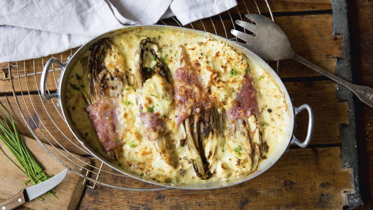 Keto-Gratin mit Chicorée in einer Auflaufform auf Holzuntergrund, Küchenutensilien drum herum.