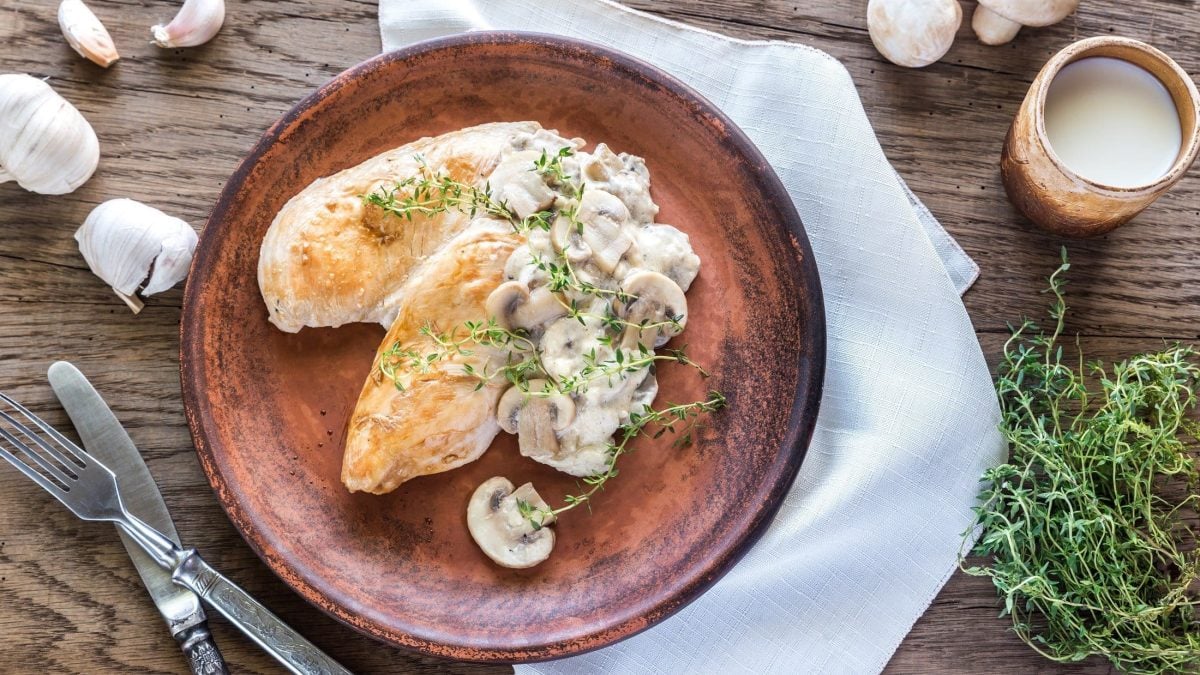 Draufsicht: Ein brauner Teller mit Knoblauch-Hühnchen in Sahne-Sauce liegt auf einem weißen Tuch, dass auf einer Holzplatte liegt. Drum herum sind ein Besteck und frischer Thymian.