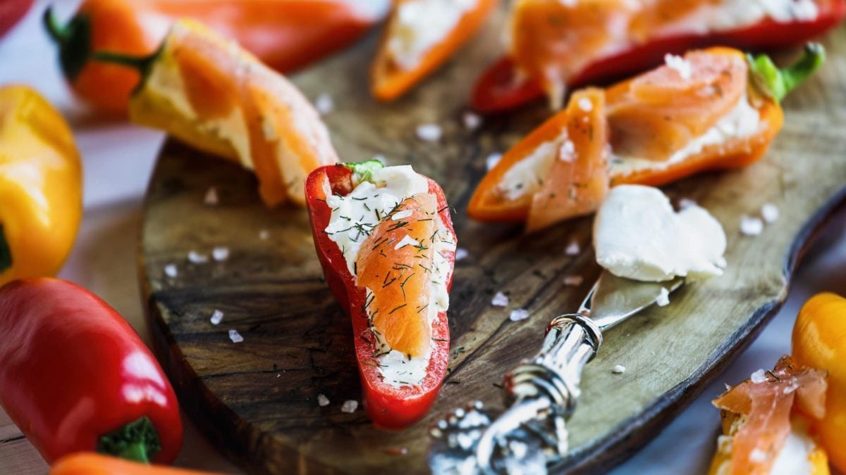 Draufsicht, schräg: Lachs-Häppchen in einer halbierten roten Spitzpaprika auf einem Holzbrettchen. Daneben liegt ein silbernen Messer.