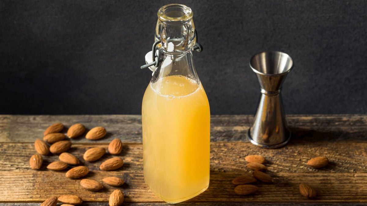 Eine Flasche mit dem Mandelsirup auf einem dunklen Holztisch mit Mandeln und einem Barmaß. Alles von der Seite fotografiert vor schwarzem Hintergrund.