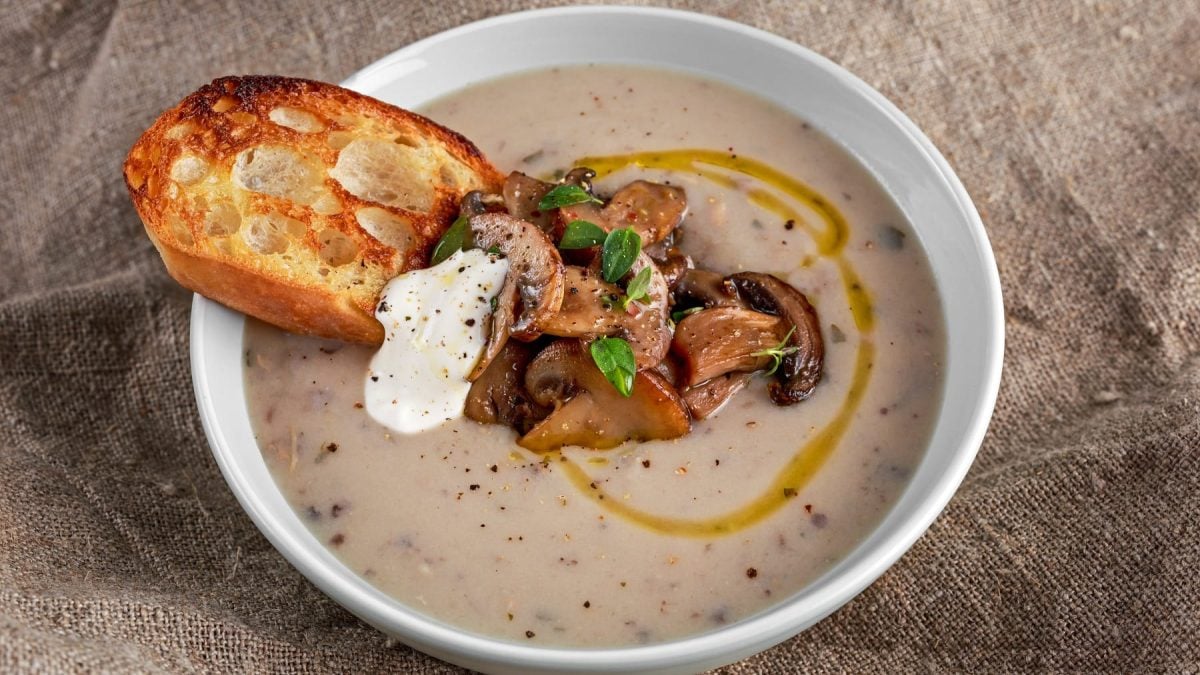 Eine weiße Schüssel mit der Maronen-Pilzcremesuppe auf einem beigen Leinentuch. In der Mitte der Suppe Pilze, Crème fraîche und geröstetes Brot.