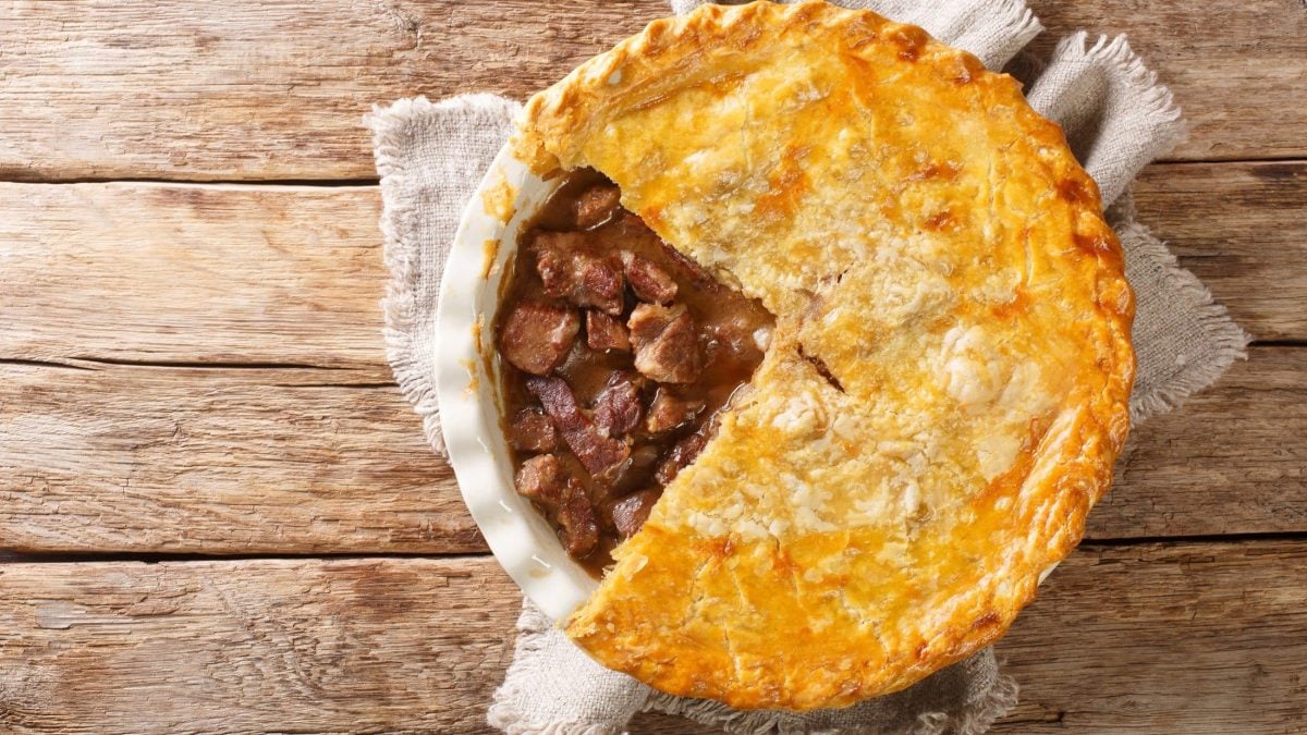 Draufsicht: Auf hellem Holz liegt eine weiße Auflaufform mit einem Meat Pie. Es handelt sich um einen Fleischkuchen. Der Meat Pie ist angeschnitten. Man sieht das gebratene Rindfleisch unter der Teighülle.