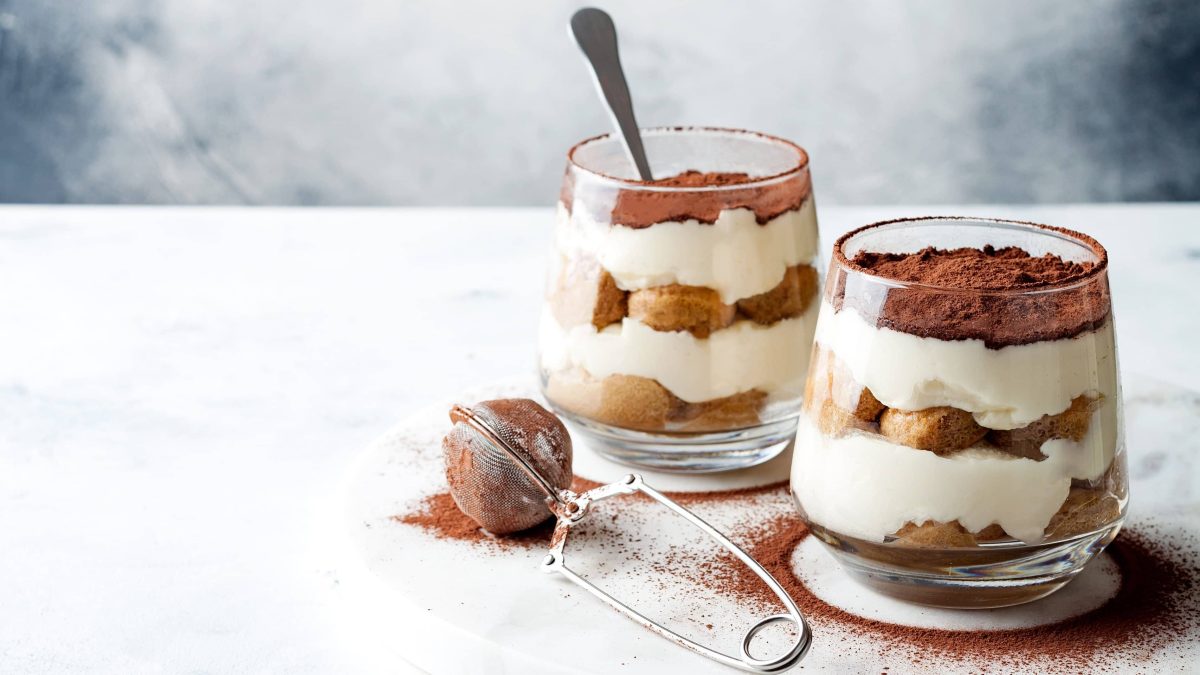 Milchmädchen-Tiramisu geschichtet in zwei Gläsern, bestreut mit Kakao, auf weißem Untergrund.