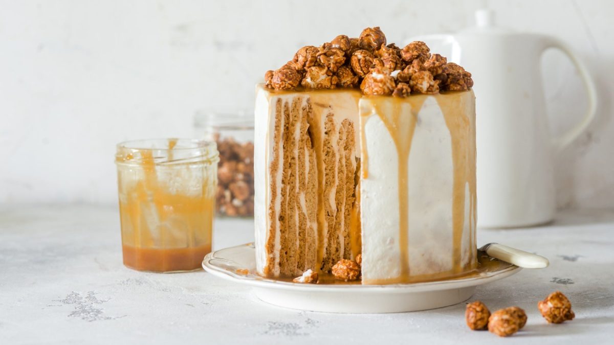 Ein Nusskuchen mit Karamell auf einem weißen Servierteller auf einem weißen Hintergrund. Links davon ein Glas mit Karamellsauce und dahinter eine Karaffe aus weißem Porzellan.