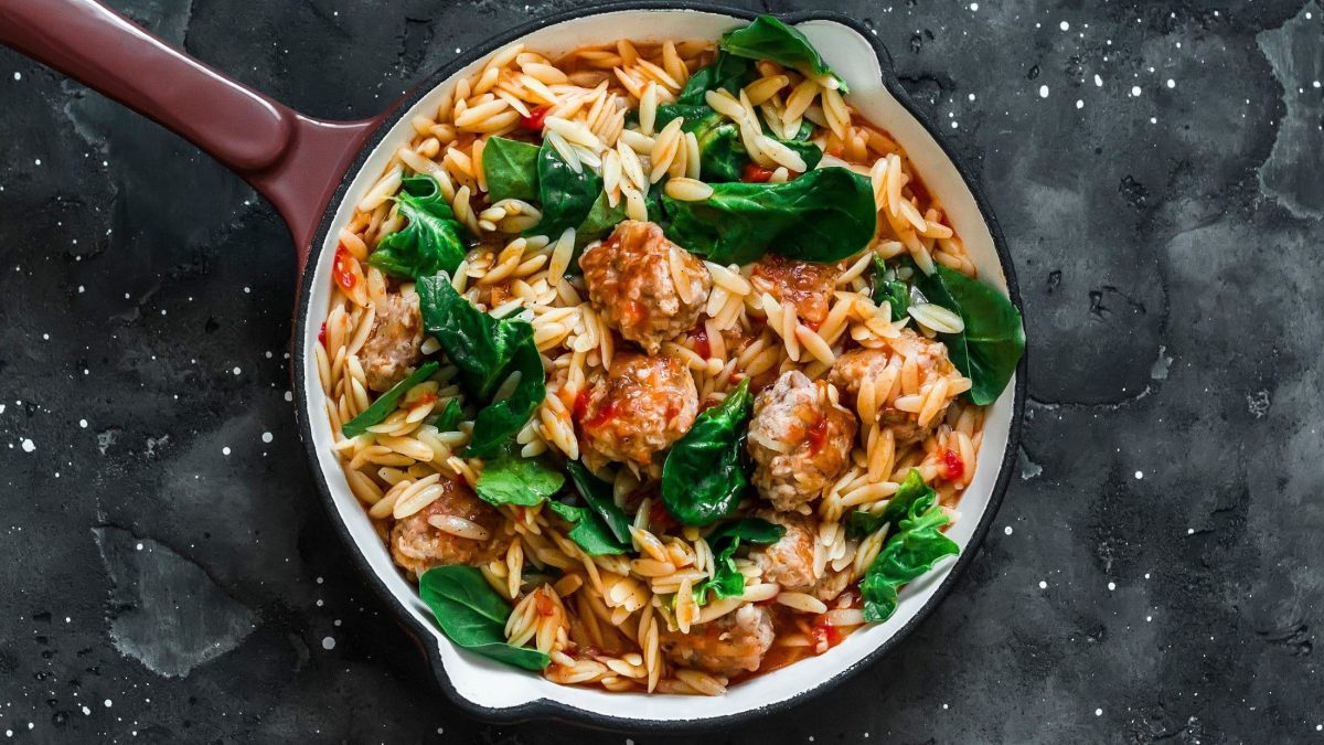 Draufblick: Ein Topf Orzo-Pasta mit Fleischbällchen auf dunklem Untergrund.