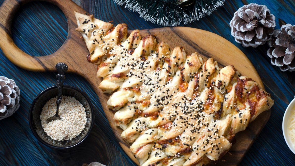 Draufsicht: Auf blauem Untergrund und einem Holzbrettchen liegt ein Puff Pastry Christmas Tree. Das ist ein Weihnachtssbaum aus Blätterteig.