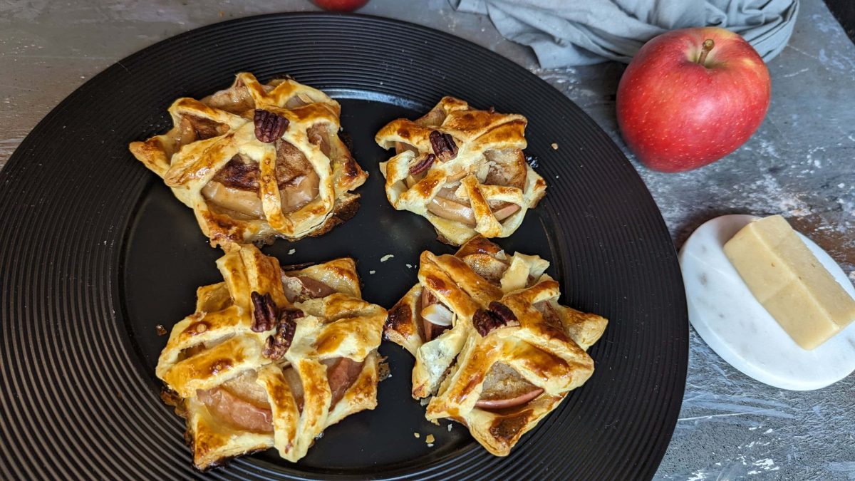 Vier Stücke Apfel-Marzipan-Plunder auf einem schwarzen Teller. Daneben Marzipan und ein roter Apfel. Alles auf einem grauen Tisch von schräg oben fotografiert.
