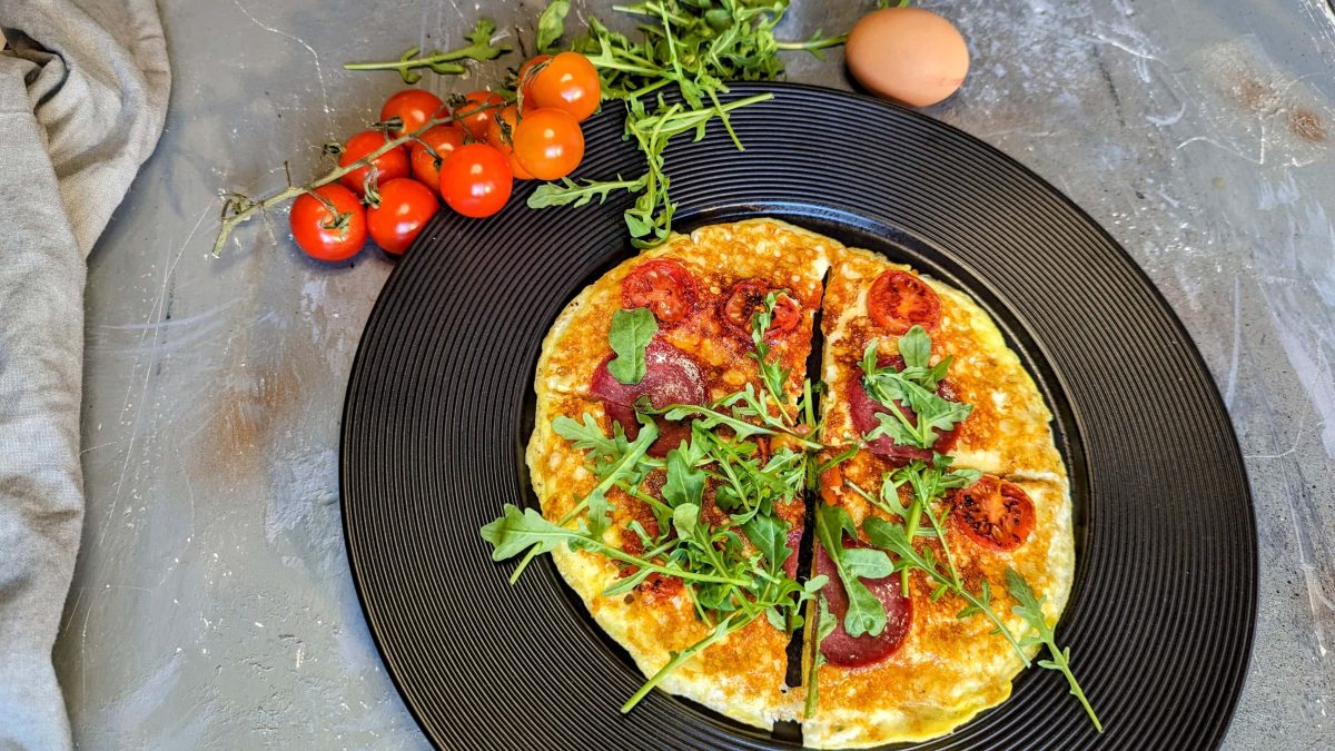 Ein schwarzer telle rmit der Frühstückspizza auf grauemj Untegrrund. Daneben Kirschtomaten, Ei und Rucola. Alles direkt von oben fotografiert.