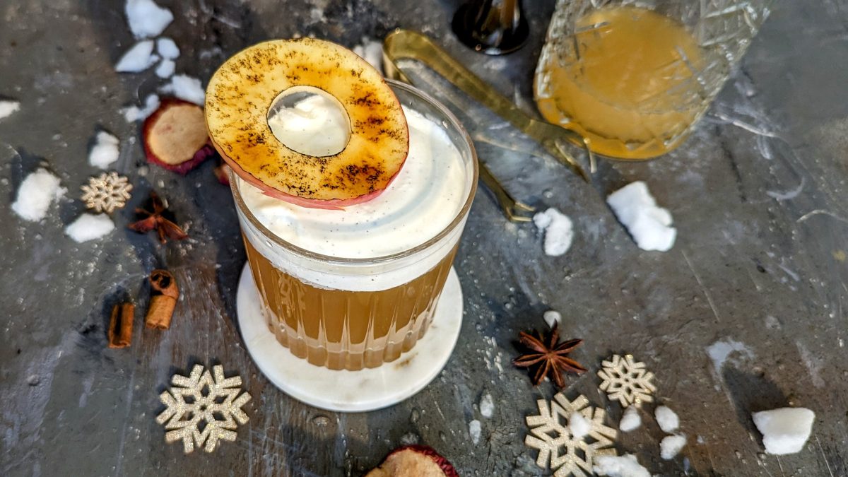 Ein Glas mit dem Old Fashioned Bratapfel auf grauem Untergrund. Daneben weihnachtliche Deko.