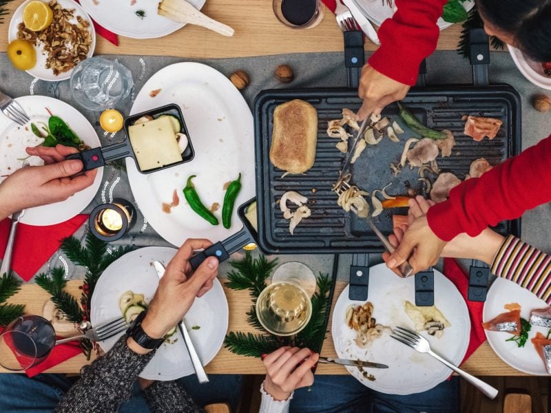 Grauer Holzuntergrund mit Raclette-Pfännchen und verschiedenen Zutaten.