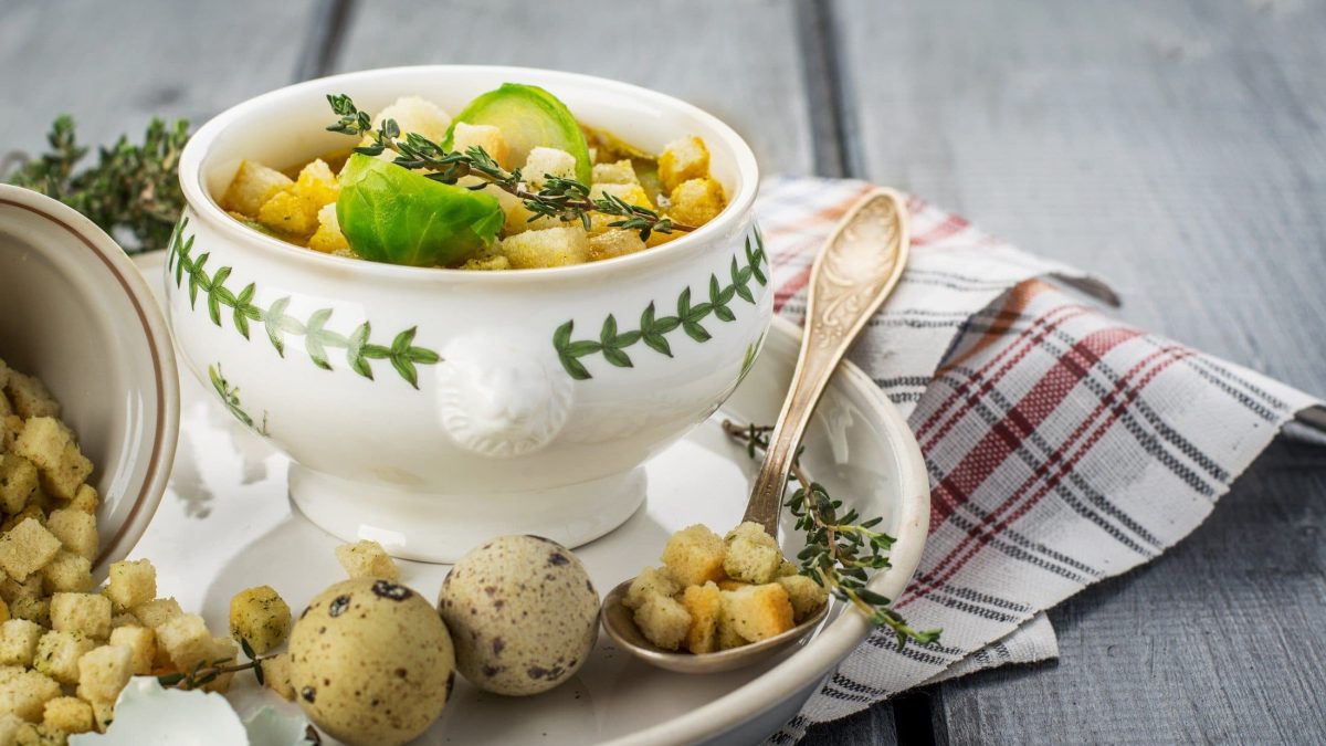 Frontal: Eine kleine Suppenschüssel mit einer Rosenkohl-Croûton-Suppe mit Wachtelei steht auf einem Teller und der auf einem Untergrund aus Holz. Drum herum liegen frischer Rosenkohl und Thymianzweige.