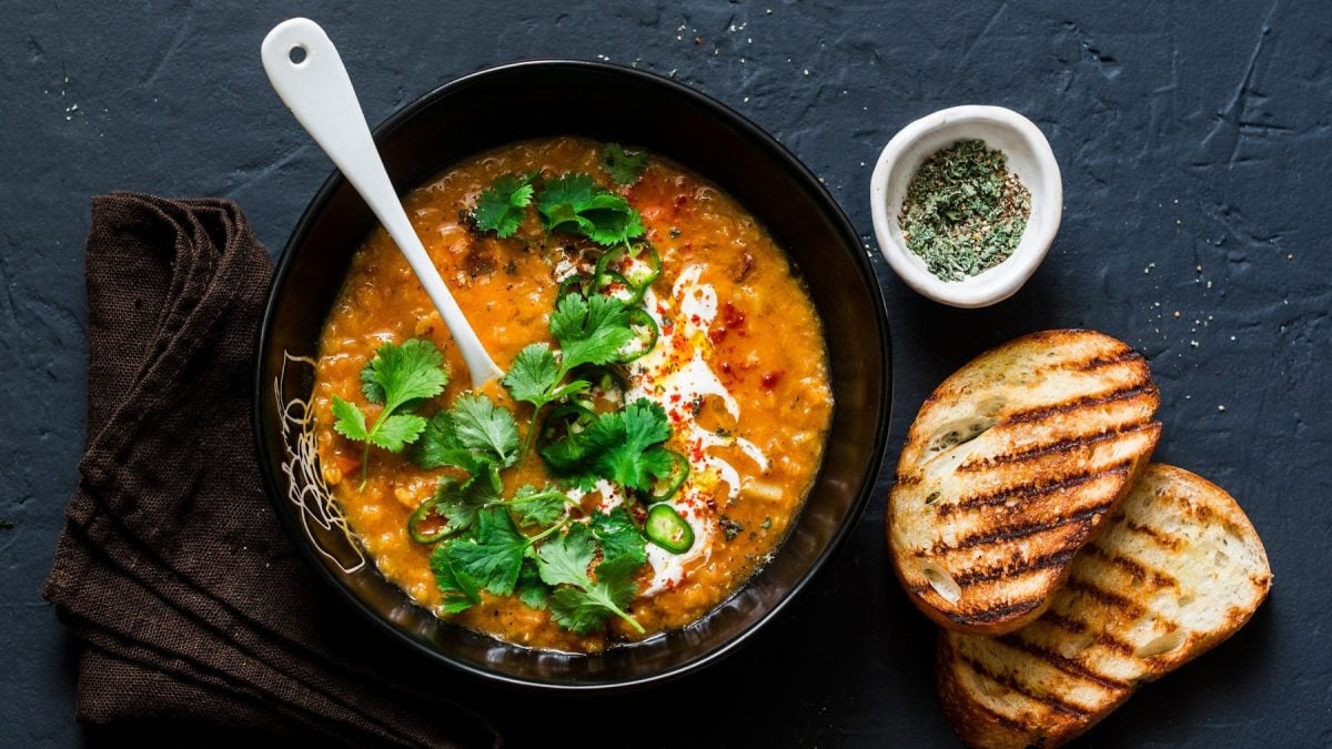 Eine schwarze Schüssel mit der Linsensuppe auf schwarzem Untergrund mit zwei Scheiben geröstetem Weißbrot und einem braunen Tuch. Alles direkt von oben fotografiert.