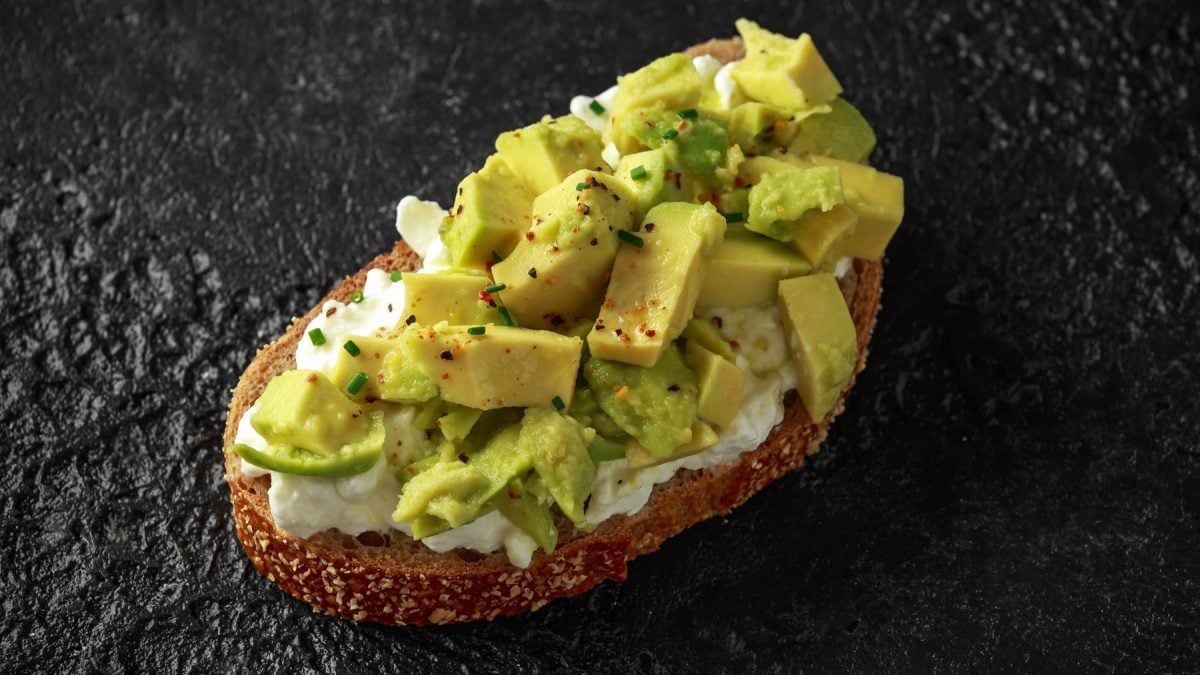 Ein belegtes Brot mit Avocado und körnigem Frischkäse auf dunklem Untergrund von schräg oben nah fotografiert.