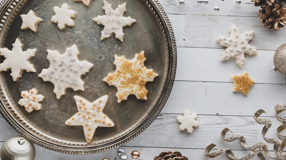Draufsicht Selbst gebackene Vanillesterne mit einer Schicht weißem Zuckerguss und goldenen und silbernen Deko-Elementen liegen auf einem silbernen Tablett, dass auf einer weißen Holzplatte liegt.