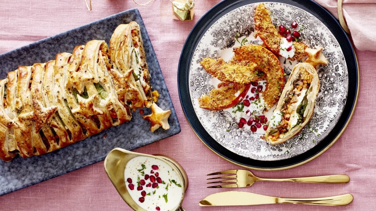 Veggiebraten "Wellington" mit Knusperkürbis auf rosarotem Hintergrund. Rechts davon ein weißer Teller angerichtet mit einer Portion. Unten ein Schälchen mit Dip und rechts davon goldenes Besteck.