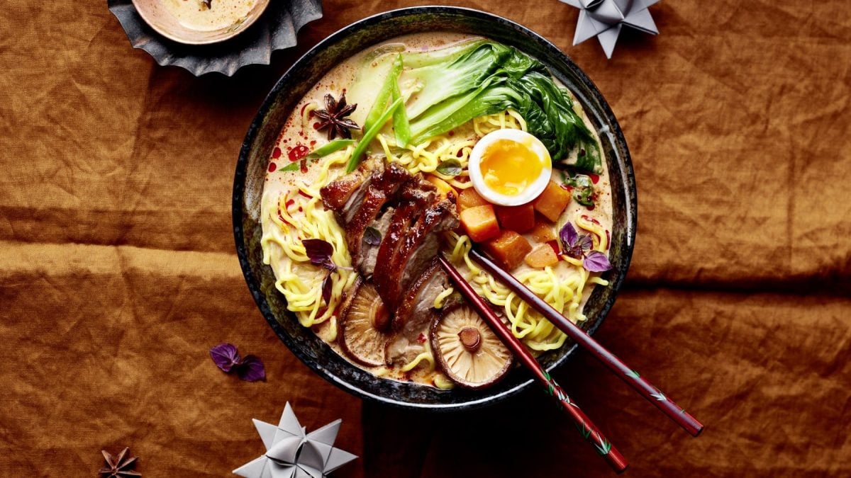 Eine Bowl mit Weihnachtsramen Eat-Club-Style auf hölzernem Hintergrund. Oben ein Teelicht und zwei Geschenkpapierschleifen und Gewürze verteilt auf dem Hintergrund.