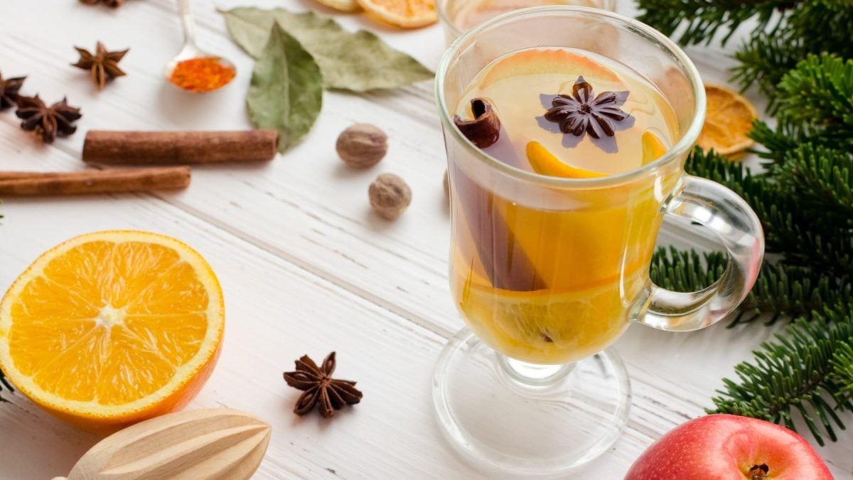 Ein Glas mit dem weißen Glühwein auf einem hellen Holztisch mit allerlei Gewürzen und winterlichen Zutaten.