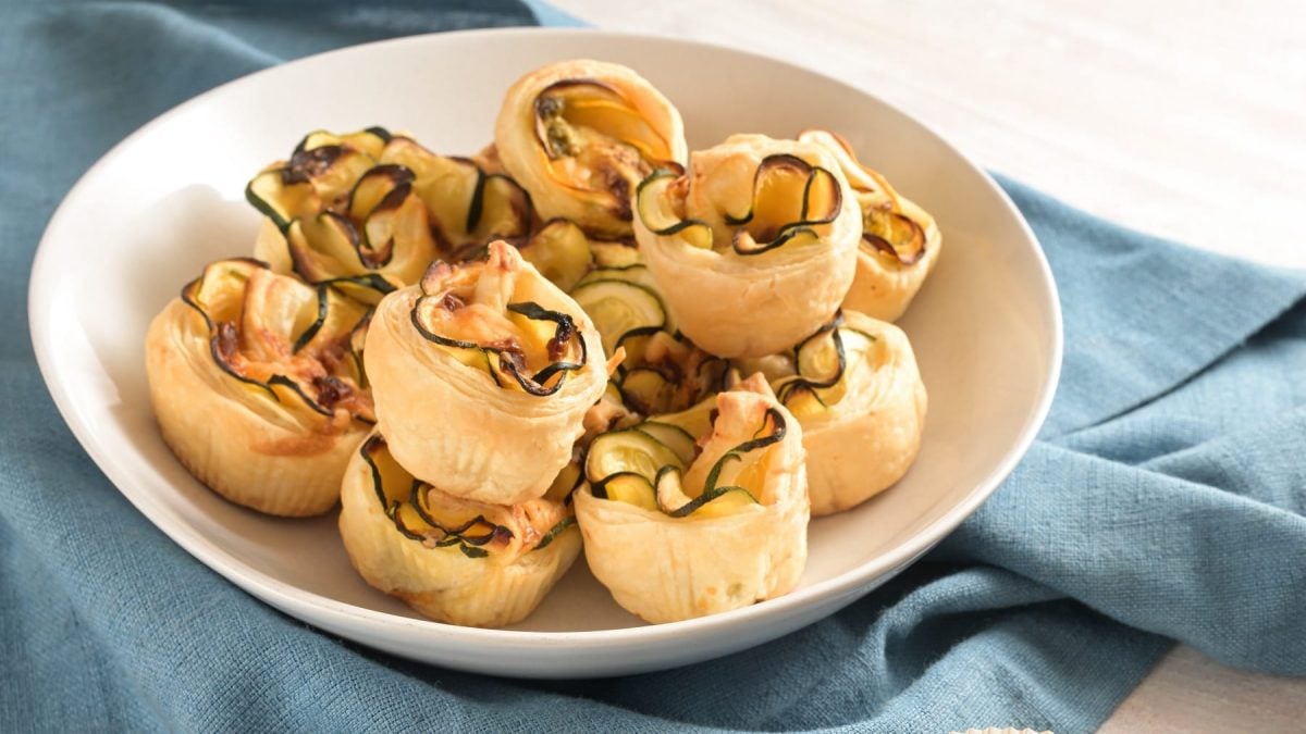 Die Zucchini-Feta-Rosen auf einem weißen Teller und blauem Tuch. Alles schräg von der Seite fotografiert.