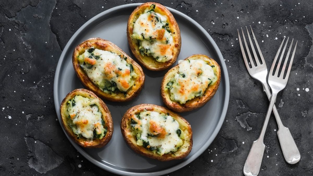 Mehrere Kartoffelhälften mit Spinat und Käse überbacken auf einem grauen Teller und dunklem Untergrund.