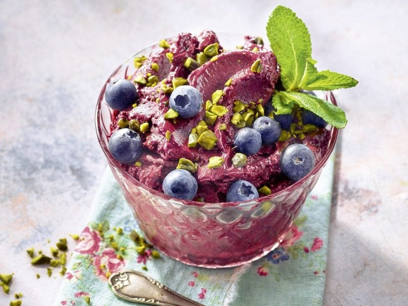Ein Dessertglas mit der Blaubeer-Bananen-Nice-Cream auf einem grauen Tisch mit hellblauer Serviette. Alles nah und von schräg oben fotografiert.