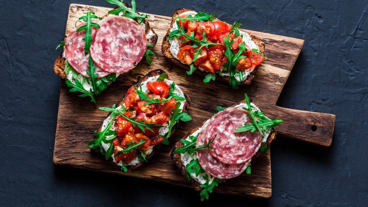 Draufsicht: Dunkler Untergrund, ein Holzbrett mit Bruschetta mit Fenchelsalami. Zwei sind zudem mit Tomatenwürfeln belegt.