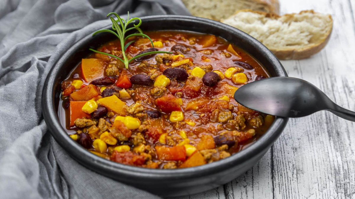Eine schwarze Schale mit dem Chili con Carne auf einem grauen Holztisch mit grauer Tischdecke auf der linken Seite. Alles von schräg oben fotografiert.